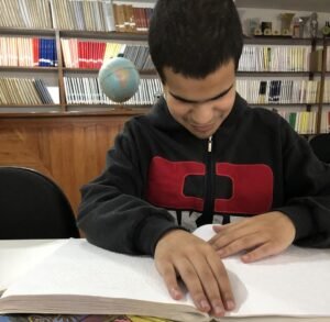 #PraTodosVerem Em uma biblioteca um jovem lê um livro em braille sobre uma mesa. Ao fundo, livros em prateleiras.